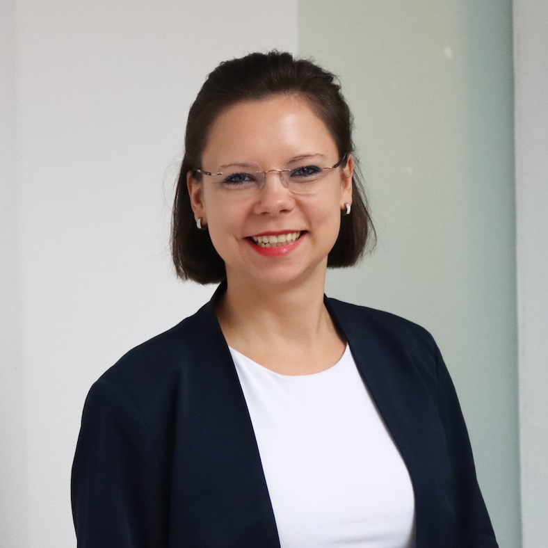 Ein Porträt von Sarah Jasiok. Ihr dunkelbraunes Haar ist etwas kürzer als mittellang. Sie trägt einen dunkelblauen Blazer, darunter ein weißes T-Shirt und eine rechteckige Brille. Sie lächelt fröhlich in die Kamera.